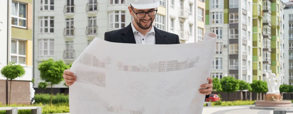 Professional architect in white hardhat holding construction projects on paper, looking and smiling. Man standing near new apartment buildings. Concept of architecture, construction, engineering.