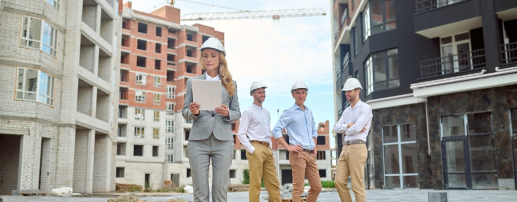Examination. Serious young adult woman in business gray suit in heels wearing safety helmet looking at document and smiling three men waiting behind at construction site